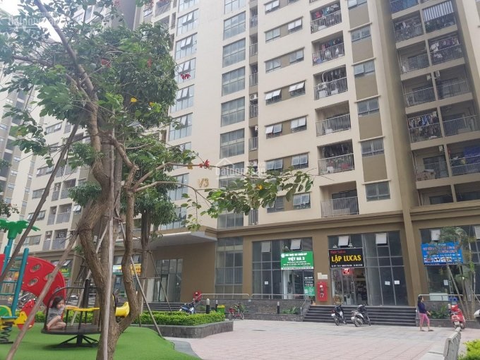 Ảnh chụp từ dưới sân một tòa nhà chung cư, cây cối, người, xe máy.