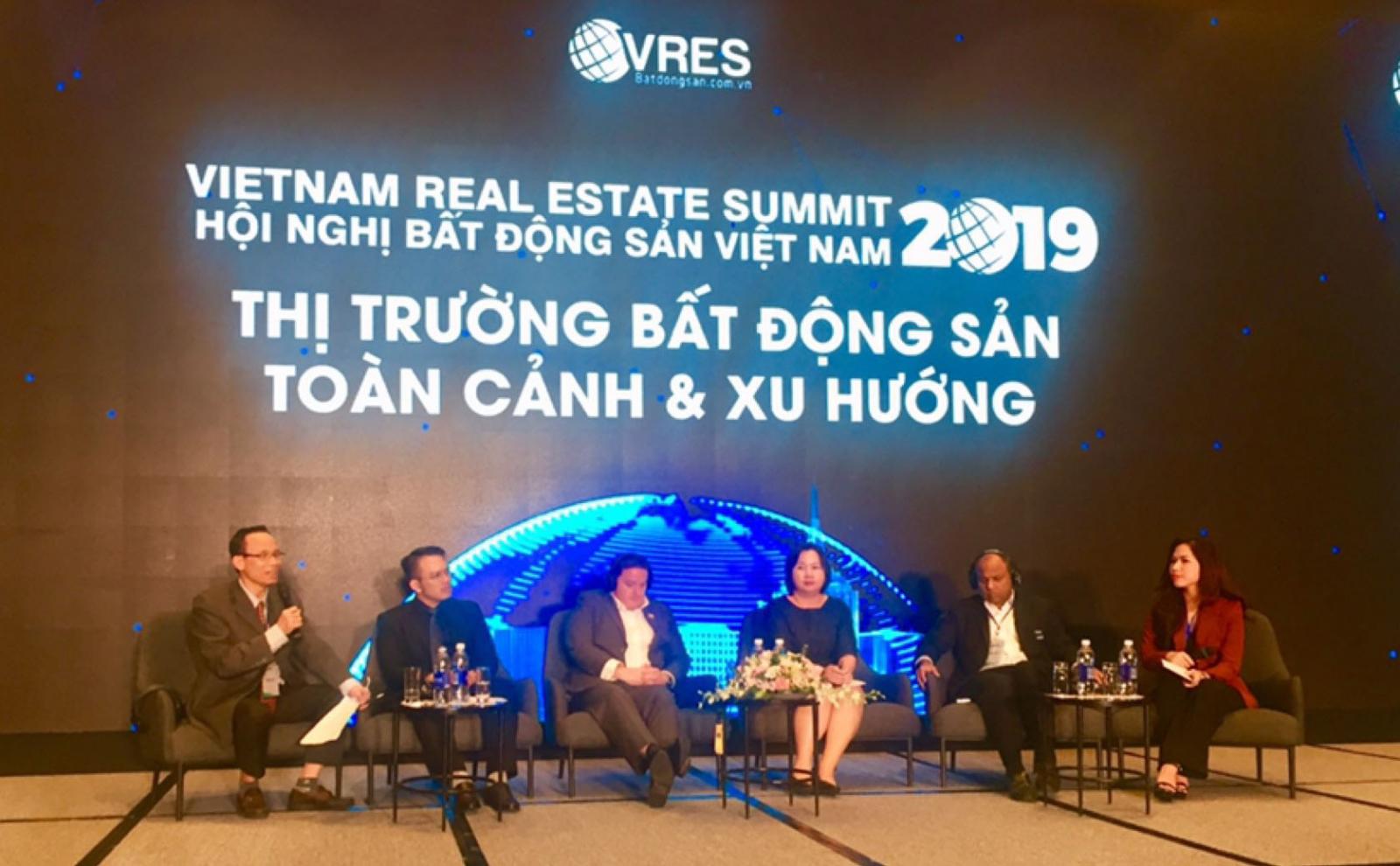 Khu vực sân khấu có phông nền tối, các dòng chữ màu trắng sáng, bên dưới là một dãy người ngồi trên ghế.
