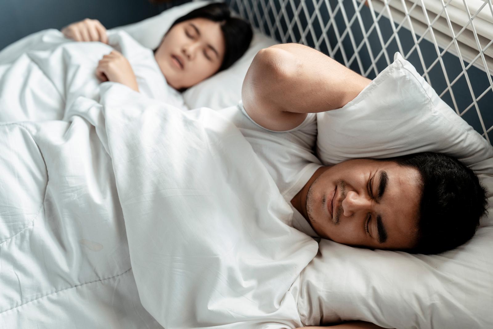 người đàn ông cau có, khó chịu  vì tiếng ồn khi đang nằm ngủ cạnh vợ