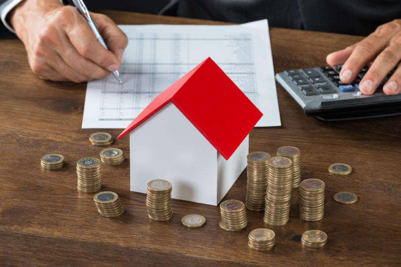 Bàn tay cầm bút ghi trên giấy, mô hình ngôi nhà, đồng xu và chiếc máy tính cầm tay