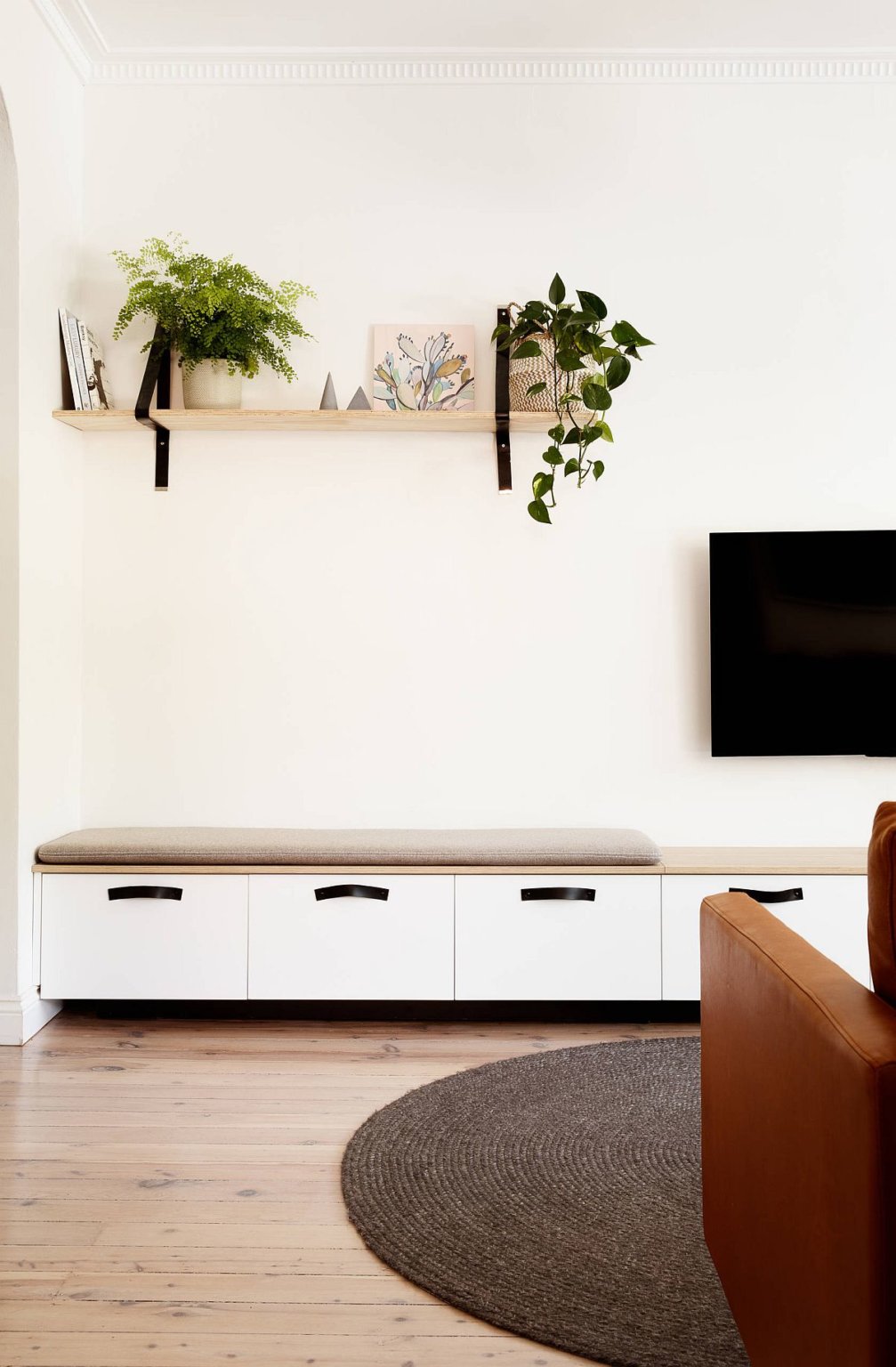 H&igrave;nh ảnh một g&oacute;c ph&ograve;ng kh&aacute;ch nhỏ với tường m&agrave;u trắng l&agrave;m nền cho kệ gỗ, c&acirc;y xanh, sofa n&acirc;u da b&ograve; nổi bật hơn