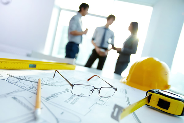 Hình ảnh cận cảnh bản vẽ thiết kế nhà và mũ bảo hộ màu vàng của thợ thi công, cạnh đó là 3 người đang trao đổi 