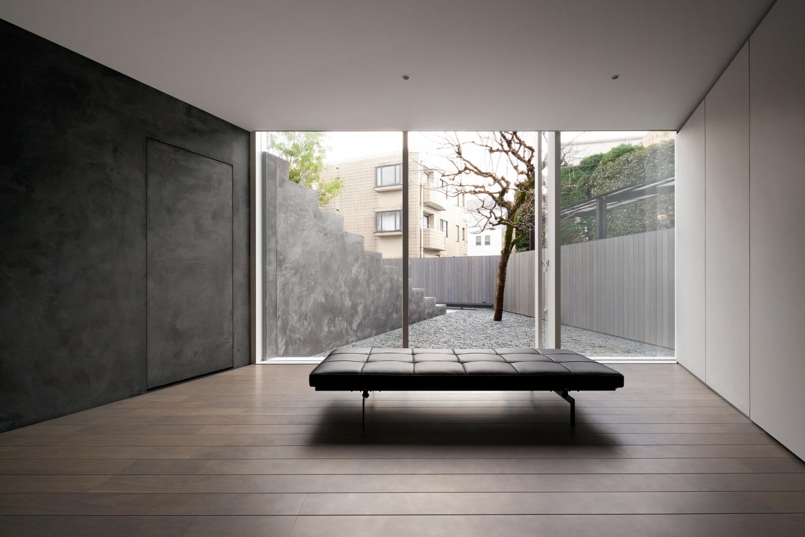 Hình ảnh một căn phòng tối giản với tường, trần sơn trắng, sofa giường đặt ở trung tâm, cửa kính mở ra sân vườn rải sỏi