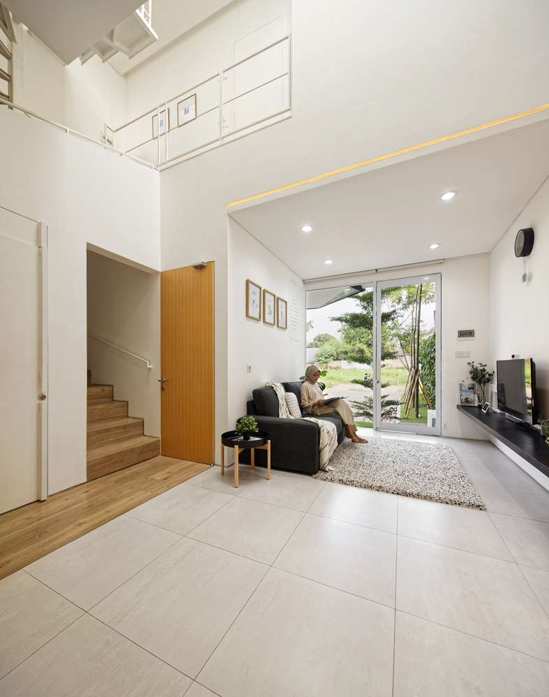 hình ảnh toàn cảnh phòng khách nhà 2 tầng với hình ảnh cô gái đang ngồi trên sofa xám đen, thảm trải sàn êm ái, cạnh sofa là bàn trà tròn nhỏ màu đen, đối diện là kệ tivi cùng màu gắn trực tiếp lên tường