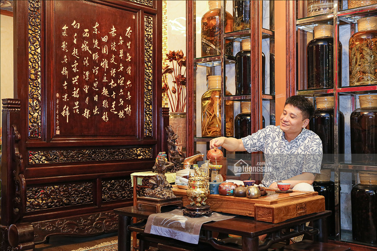 Người đ&agrave;n &ocirc;ng đang r&oacute;t tr&agrave;, xung quanh l&agrave; ph&ograve;ng kh&aacute;ch b&agrave;y nhiều đồ gỗ mỹ nghệ