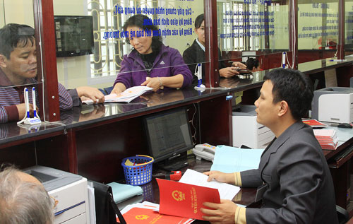 3 người đang đứng trong quầy giao dịch, tr&ecirc;n b&agrave;n l&agrave;m việc c&oacute; giấy tờ v&agrave; một cuốn sổ đỏ