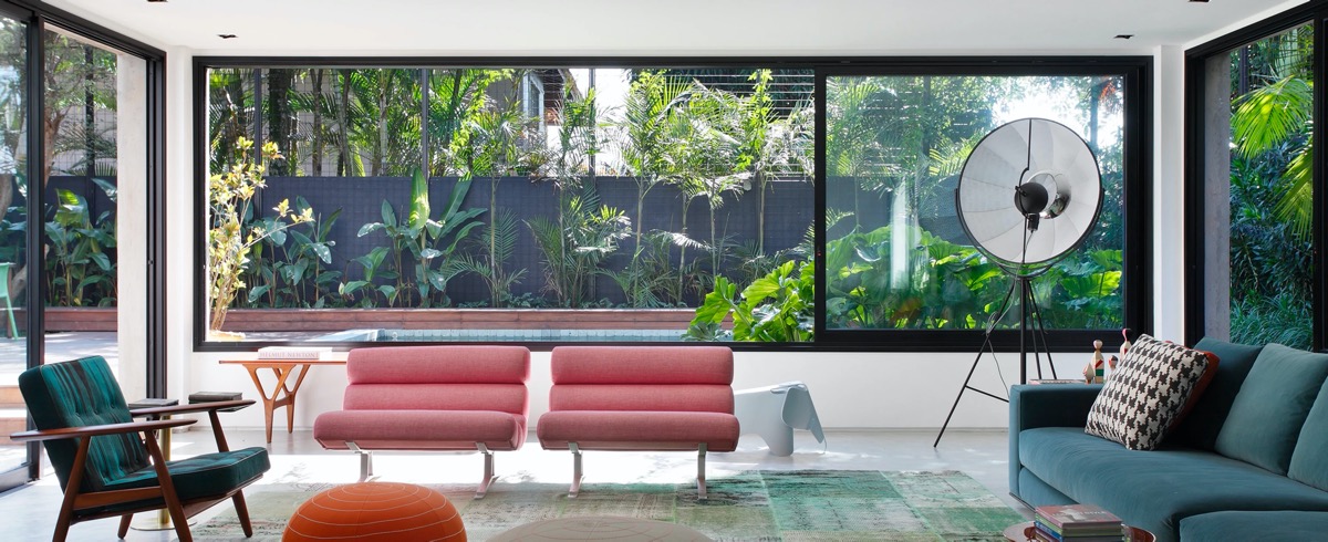 h&igrave;nh ảnh ph&ograve;ng kh&aacute;ch ng&ocirc;i nh&agrave; miền nhiệt đới với sofa m&agrave;u xanh ngọc, ghế thư gi&atilde;n m&agrave;u hồng, đ&ocirc;n ngồi m&agrave;u cam, cửa sổ k&iacute;nh lớn mở ra s&acirc;n vườn trồng nhiều c&acirc;y xanh