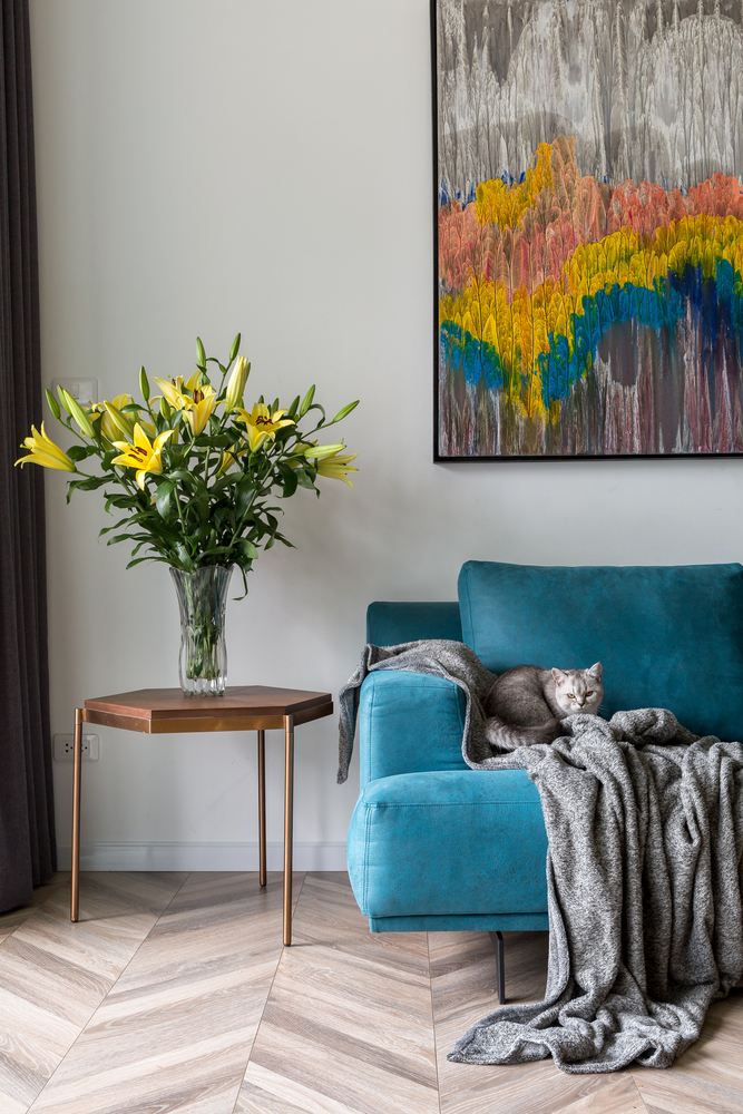 h&igrave;nh ảnh g&oacute;c ph&ograve;ng kh&aacute;ch hiện đại với sofa nhung m&agrave;u xanh dương, tranh tường nghệ thuật, b&agrave;n phụ kim loại s&aacute;ng b&oacute;ng, tr&ecirc;n b&agrave;n đặt b&igrave;nh hoa ly m&agrave;u v&agrave;ng