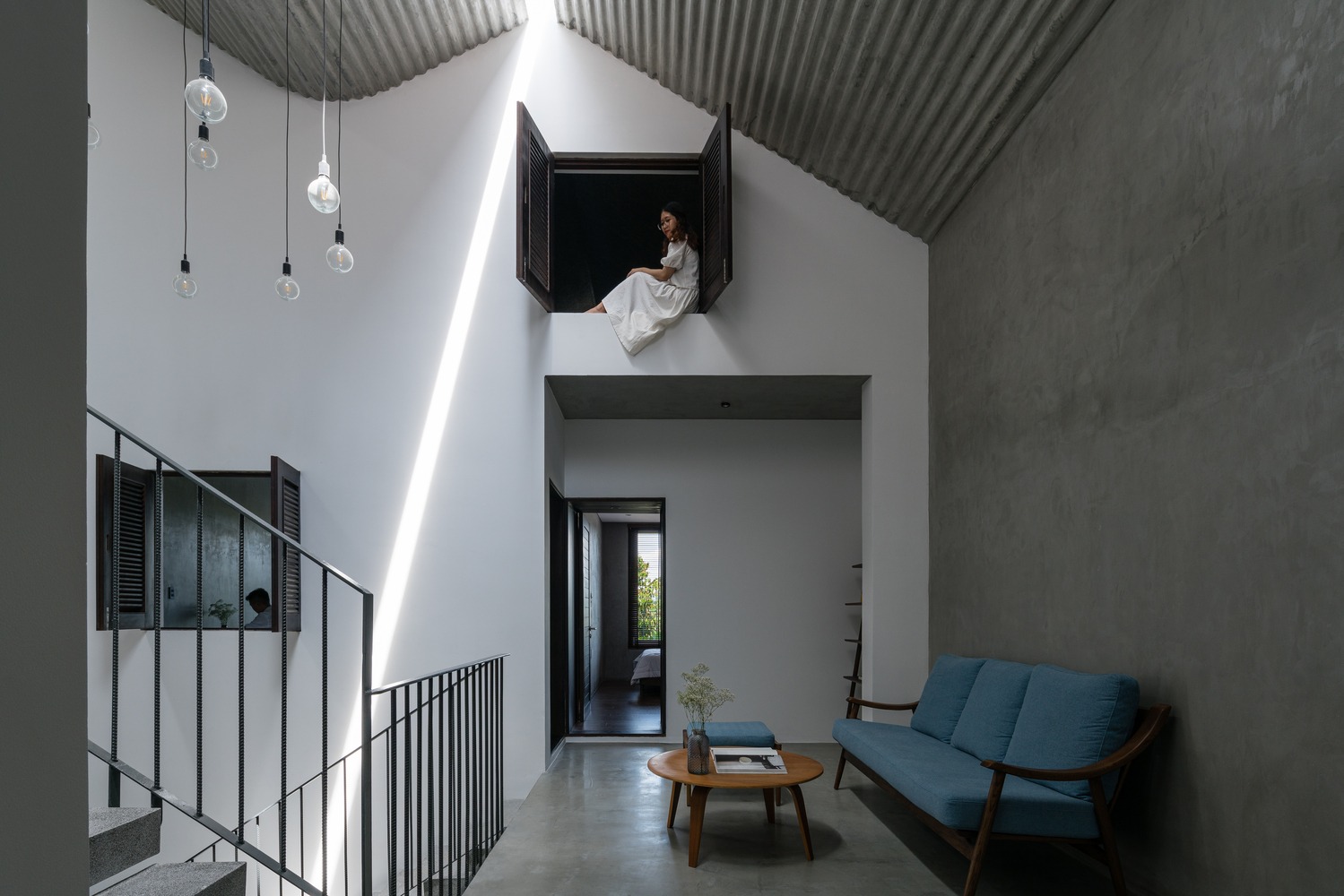 h&igrave;nh ảnh c&ocirc; g&aacute;i ngồi tr&ecirc;n bậu cửa sổ, ph&aacute;i tầng dưới l&agrave; b&agrave;n gỗ, ghế sofa d&agrave;i bọc nệm m&agrave;u xanh dương, đ&egrave;n thả sợi đốt