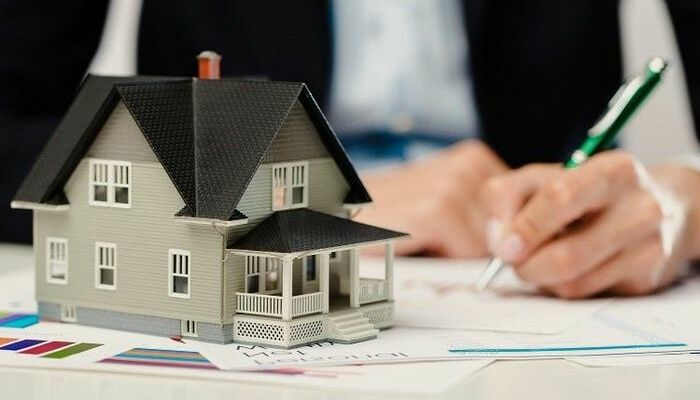 hình ảnh cận cảnh mô hình ngôi nhà với mái ngói màu đen, tường ốp gỗ xám, phía sau là hình người mặc vest đen cầm bút ký vào giấy tờ minh họa cho việc nộp tiền sử dụng đất