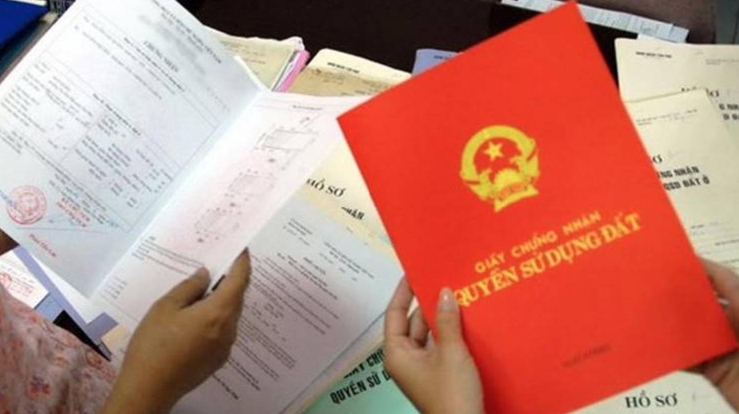 một bàn tay đang cầm sổ đỏ, xung quanh là nhiều loại giấy tờ khác