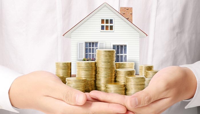 hình ảnh một người mặc sơ mi dài tay màu trắng, hai tay ôm mô hình ngôi nhà và các cọc tiền xu vàng