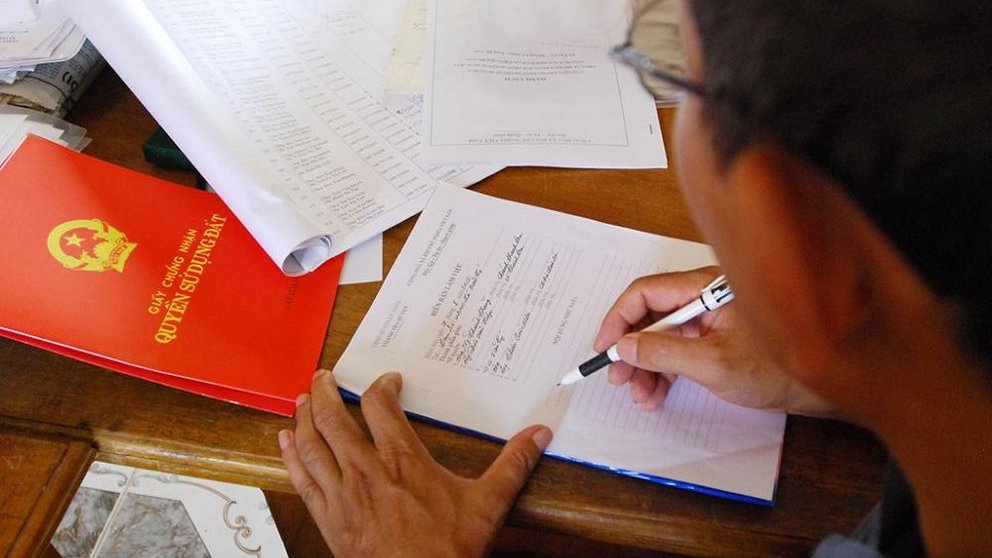 h&igrave;nh ảnh cận cảnh người đ&agrave;n &ocirc;ng luống tuổi viết v&agrave;o bản hợp đồng, trước mặt l&agrave; cuốn sổ đỏ