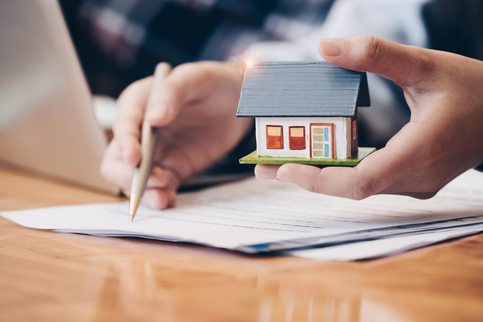 hình ảnh bàn tay cầm mô hình ngôi nhà, tay kia cầm bút ký vào giấy tờ minh họa cho thủ tục sang tên nhà đất