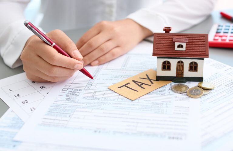hình ảnh một người cầm bút màu đỏ chuẩn bị viết, cạnh đó là mô hình ngôi nhà, máy tính, các đồng tiên xu