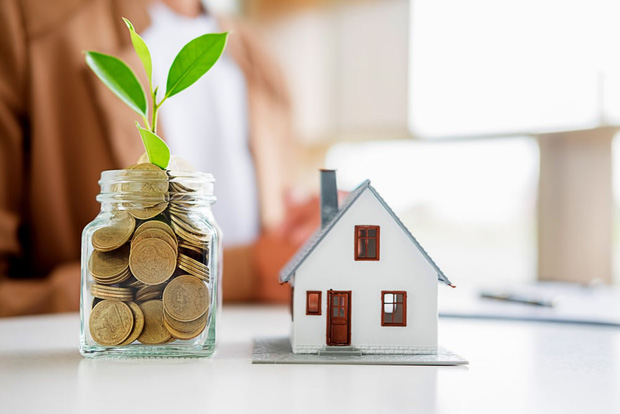 Mua chung cư trả góp mô hình ngôi nhà cạnh bình tiền xu, phía trên cắm cành lá xanh tươi