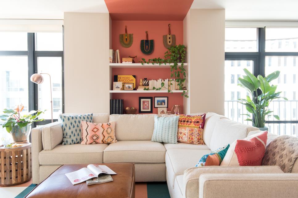 to&agrave;n cảnh ph&ograve;ng kh&aacute;ch với sofa m&agrave;u trắng sữa, mảng tường m&agrave;u cam nơi c&oacute; gi&aacute; s&aacute;ch b&agrave;y đồ trang tr&iacute;