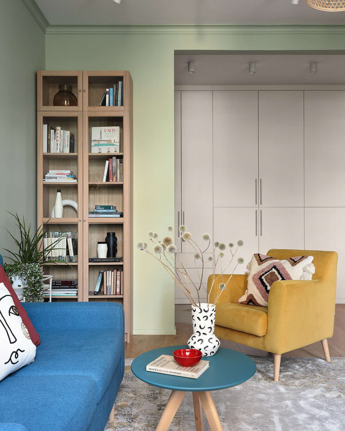 Chiếc b&igrave;nh gốm đen v&agrave; trắng li&ecirc;n kết với c&aacute;c đối t&aacute;c đơn sắc được hiển thị b&ecirc;n trong một tủ s&aacute;ch bằng gỗ, trong khi sắc xanh của c&acirc;y l&aacute; bổ sung cho chiếc ghế m&agrave;u v&agrave;ng. 