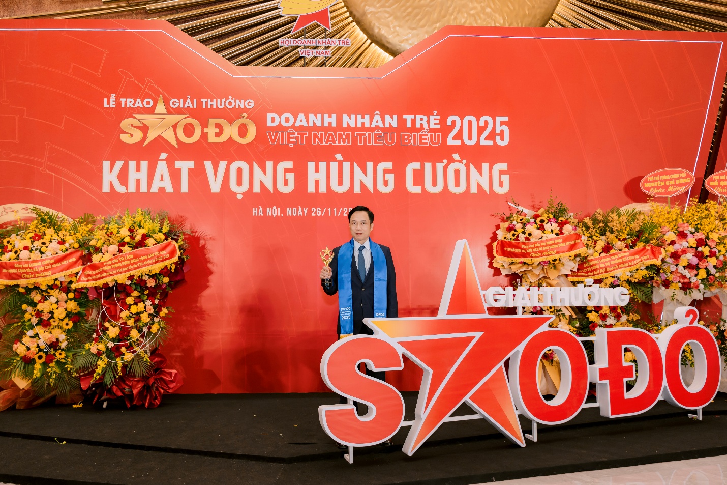 Tổng Giám đốc Dược Nam Hà được vinh danh Doanh nhân trẻ Việt Nam tiêu biểu 2025- Ảnh 2. A person standing in front of a red sign Description automatically generated